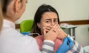 Przerażona pacjentka zakrywa usta, gdy dentystka zbliża się z narzędziami. Konsekwencje nie mycia zębów na noc.