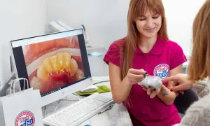 Dentystka pokazuje pacjentce model zębów, omawiając problem kamienia nazębnego szybko powracającego. Na ekranie komputera widać zbliżenie zębów z widocznym osadem.