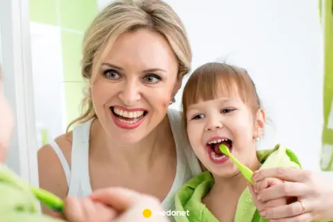 Mama uczy córkę, jak myć zęby niemowlakowi. Uśmiechnięte twarze i zielone szczoteczki.