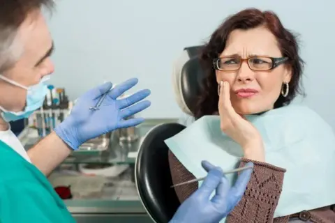 Kobieta z bólem zęba u dentysty, który pokazuje narzędzia. Wygląda na to, że czeka ją leczenie zatrucia zęba.