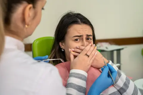 Przerażona pacjentka zakrywa usta, gdy dentystka zbliża się z narzędziami. Konsekwencje nie mycia zębów na noc.
