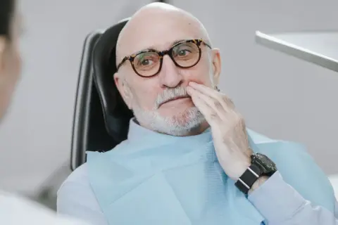 Starszy mężczyzna w okularach siedzi na fotelu dentystycznym, trzymając się za policzek. Wygląda na to, że boli go rozchwiany ząb.