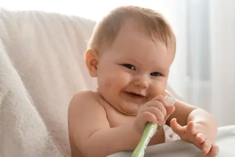 Uśmiechnięte niemowlę bawi się swoją pierwszą szczoteczką do zębów dla niemowląt, odkrywając higienę jamy ustnej.