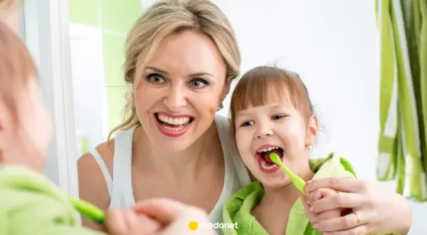 Mama uczy córkę, jak myć zęby niemowlakowi. Uśmiechnięte twarze i zielone szczoteczki.