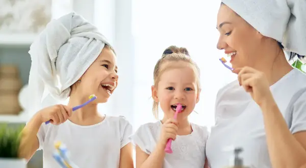 Mama z córkami myje zęby. Od kiedy myć zęby dziecku? Od pierwszego ząbka! Uśmiechnięte buzie i szczoteczki w rękach.