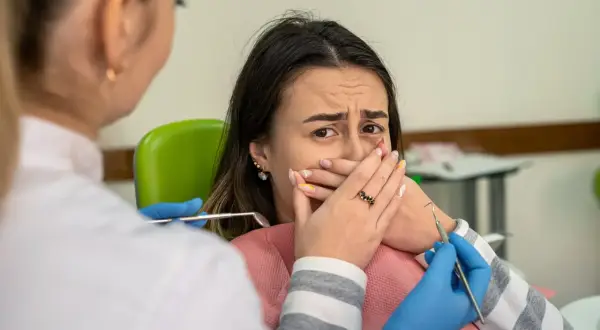 Przerażona pacjentka zakrywa usta, gdy dentystka zbliża się z narzędziami. Konsekwencje nie mycia zębów na noc.