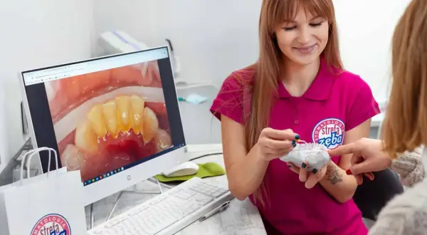 Dentystka pokazuje pacjentce model zębów, omawiając problem kamienia nazębnego szybko powracającego. Na ekranie komputera widać zbliżenie zębów z widocznym osadem.