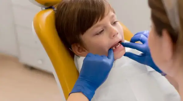 Chłopiec na żółtym fotelu dentystycznym, u którego lekarz w niebieskich rękawiczkach bada zęby, by wykluczyć choroby dziąseł u dzieci.
