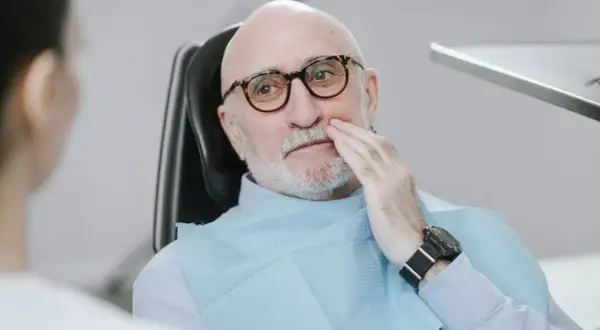 Starszy mężczyzna w okularach siedzi na fotelu dentystycznym, trzymając się za policzek. Wygląda na to, że boli go rozchwiany ząb.