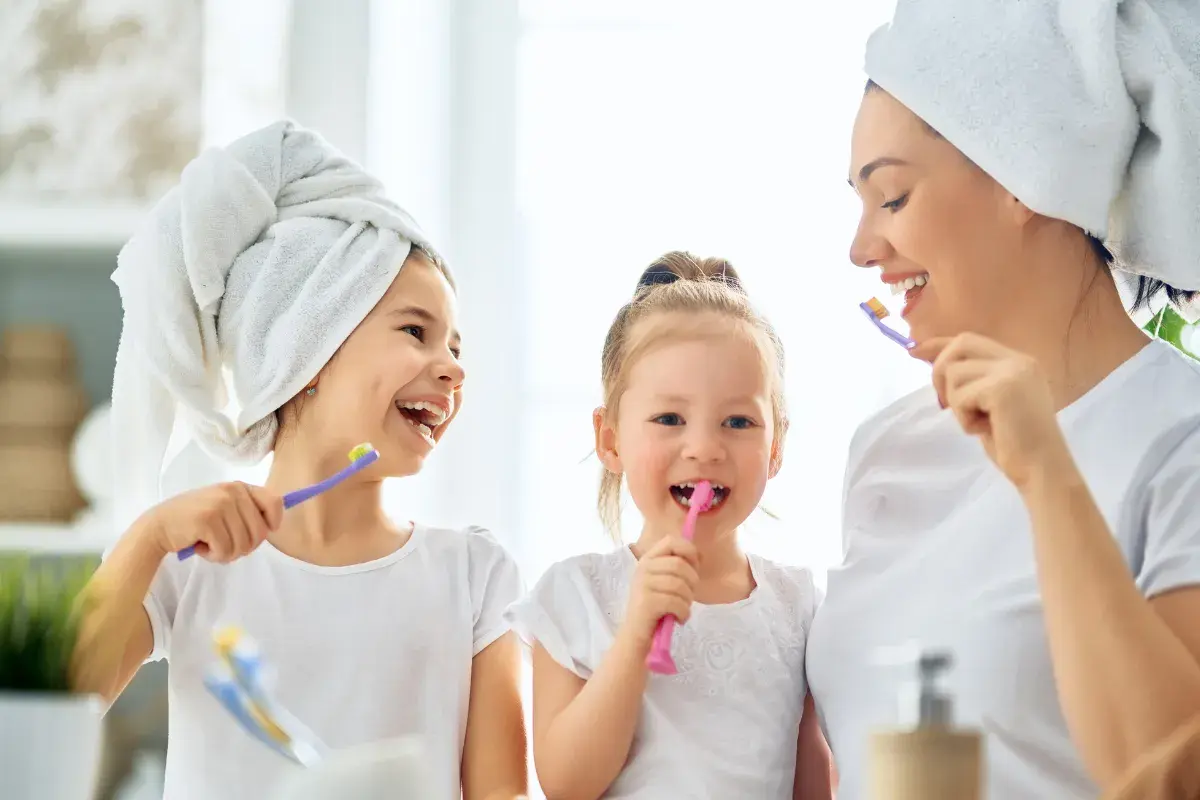 Mama z córkami myje zęby. Od kiedy myć zęby dziecku? Od pierwszego ząbka! Uśmiechnięte buzie i szczoteczki w rękach.