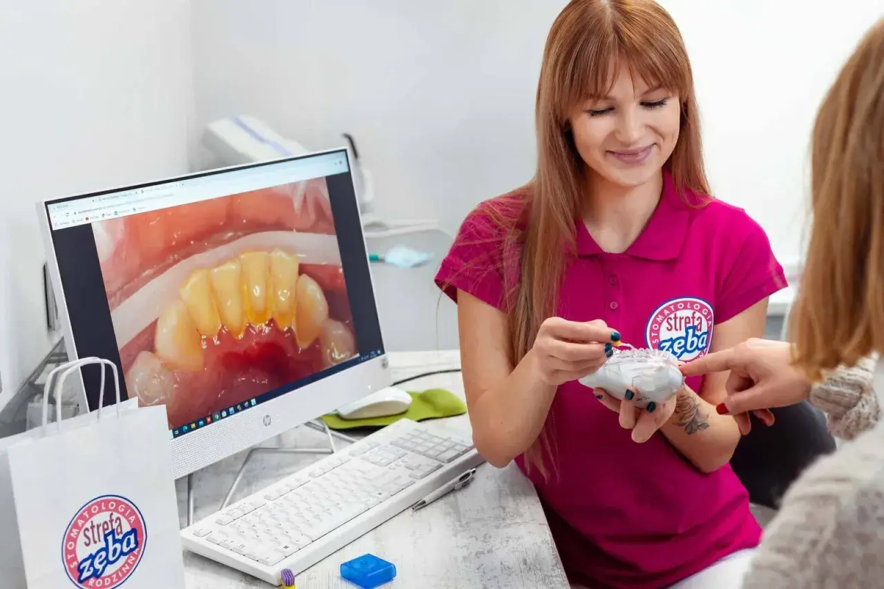 Dentystka pokazuje pacjentce model zębów, omawiając problem kamienia nazębnego szybko powracającego. Na ekranie komputera widać zbliżenie zębów z widocznym osadem.