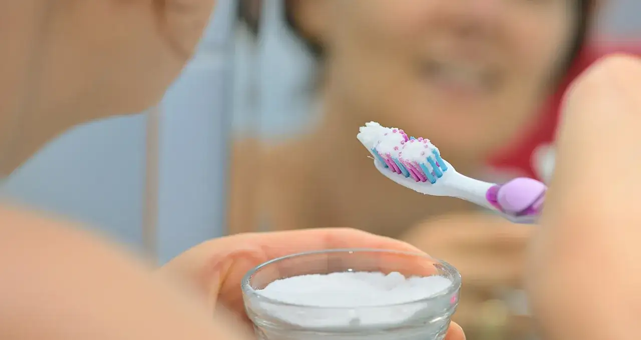 Przygotowanie do mycia zębów sodą. Na szczoteczce biały proszek, w dłoni szklana miseczka z sodą.