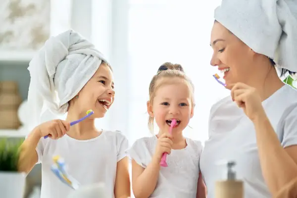 Mama z córkami myje zęby. Od kiedy myć zęby dziecku? Od pierwszego ząbka! Uśmiechnięte buzie i szczoteczki w rękach.