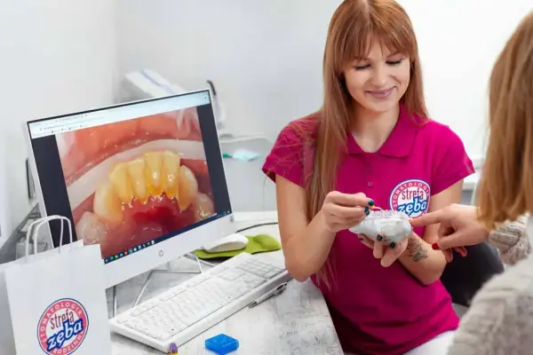 Dentystka pokazuje pacjentce model zębów, omawiając problem kamienia nazębnego szybko powracającego. Na ekranie komputera widać zbliżenie zębów z widocznym osadem.